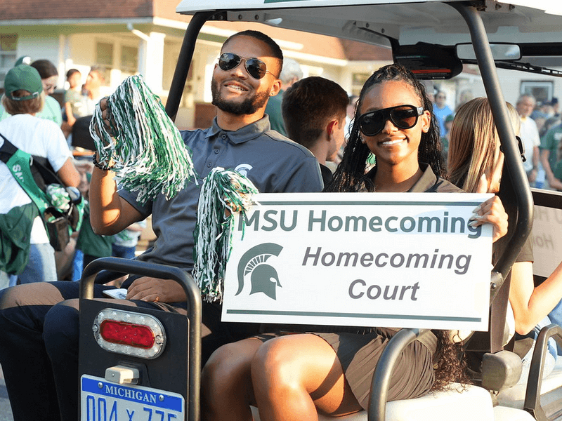 african american male and female student at homecoming parade riding in a golf cart