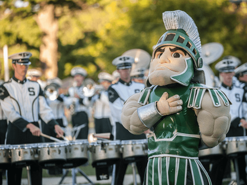 sparty standing with hand to heart with msu band playing behind