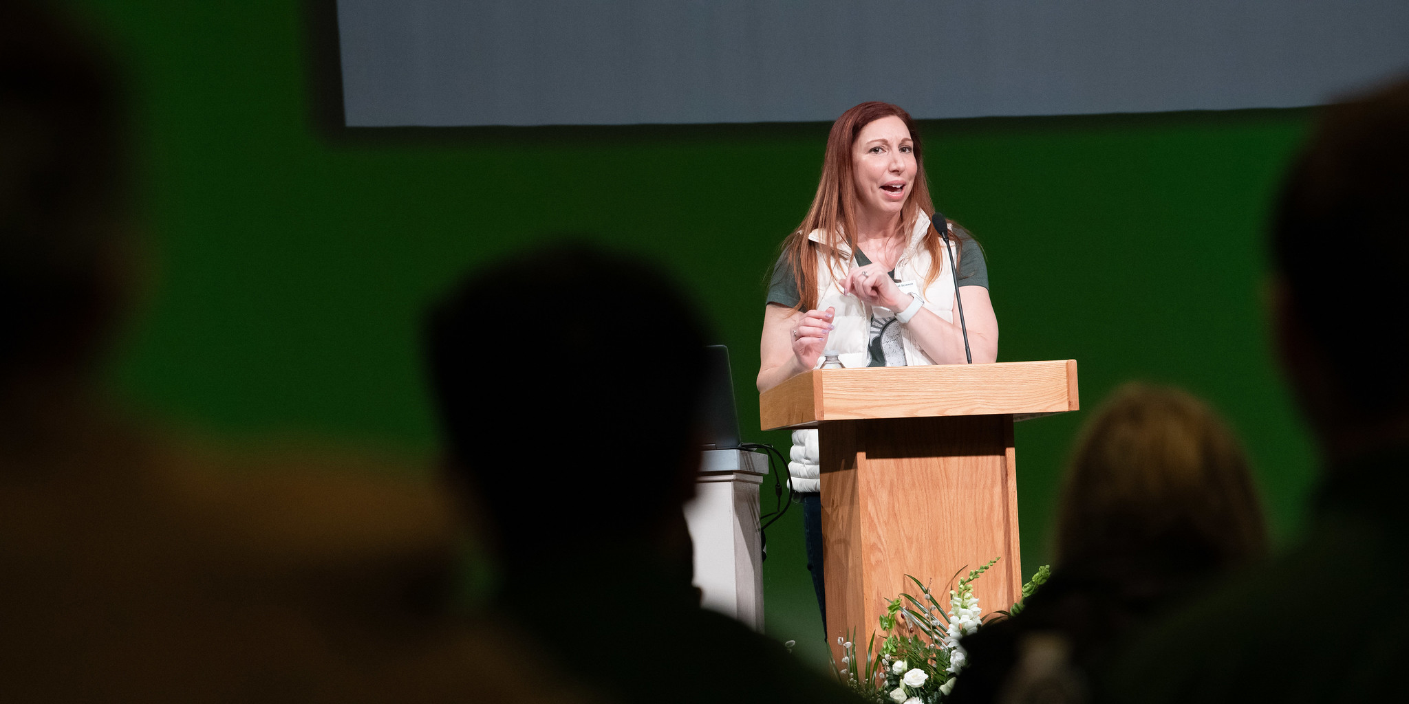A person is giving a lecture at the 2025 MSU College of Natural Science "Classes Without Quizzes" series, standing behind a wooden podium with a laptop. The background features a green and white sign that reads "CLASSES without QUIZZES." Several audience members are visible in the foreground.