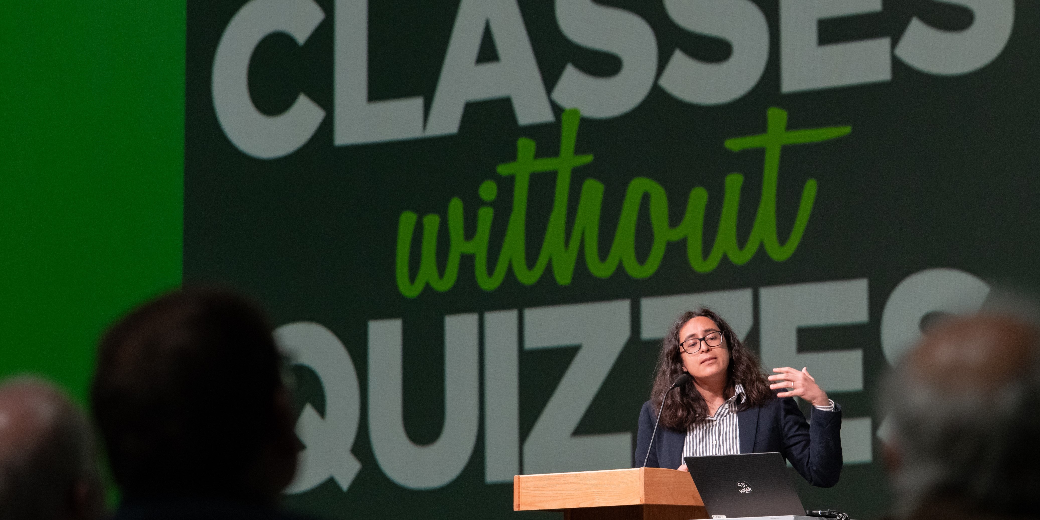 A person is giving a lecture at the 2025 MSU College of Natural Science "Classes Without Quizzes" series, standing behind a wooden podium with a laptop. The background features a green and white sign that reads "CLASSES without QUIZZES." Several audience members are visible in the foreground.