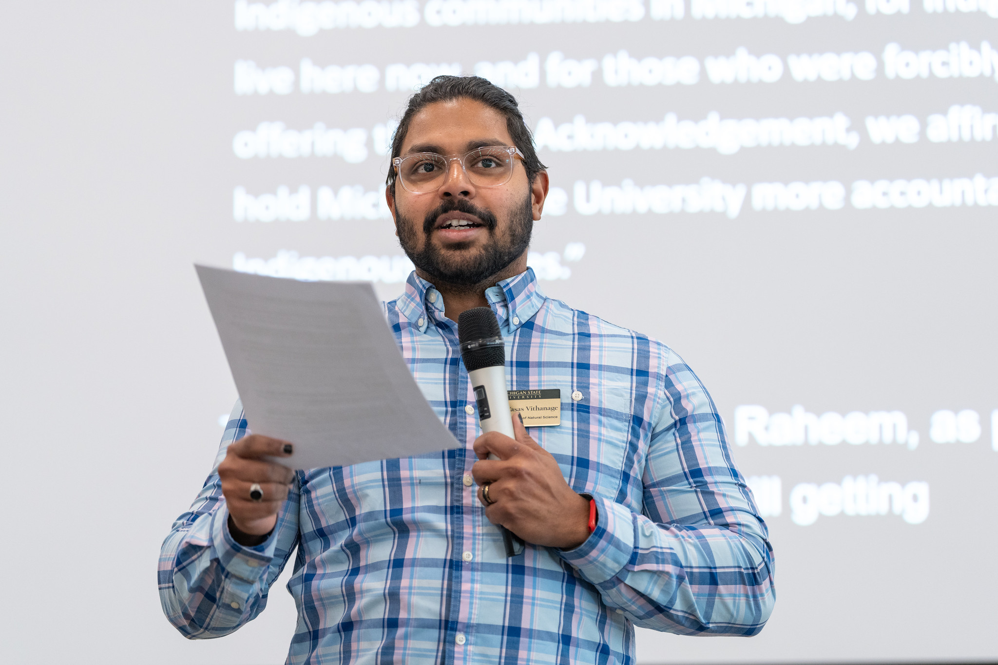 Person speaking in front of a projection screen holding a microphone and papers