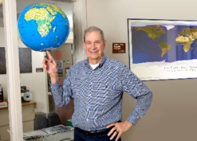Jim Tiedje standing in front of the office door to the MSU Center for Microbial Ecology with an inflatible globe balanced on his right index finger.