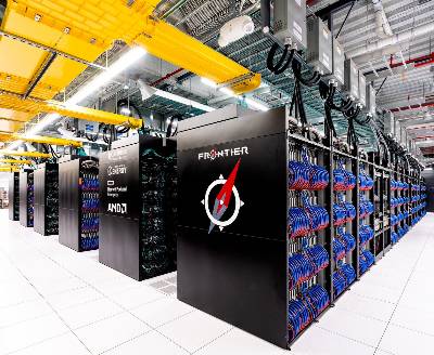 The Frontier supercomputer has a large footprint in a cavernous room at Oak Ridge National Laboratory. The supercomputer is made up of 74 supercomputing cabinets, several of which are visible in the photograph. Two of these tall black towers are labeled with text reading “Frontier,” “Oak Ridge National Laboratory, “U.S. Department of Energy,” “Hewlett Packard Enterprise,” and “AMD.” All told, the cabinets house 9,400 computing nodes, which are connected to one another with red and blue cables. 