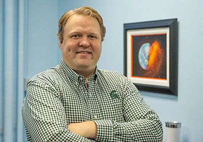 MSU planetrary scientist Seth Jacobson standing in his office with a framed astronomy picture in the background.