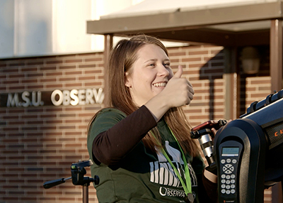 Michigan State University senior Emma Dugan gives a thumbs-up standing next to a telescope in front of the MSU Observatory. 