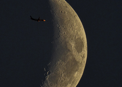 A photograph taken with the help of the MSU Observatory’s telescope shows an airplane flying in front of a waxing crescent moon. The plane is in front the dark side of the crescent. Craters and smooth areas are detailed on the crescent’s bright side. 