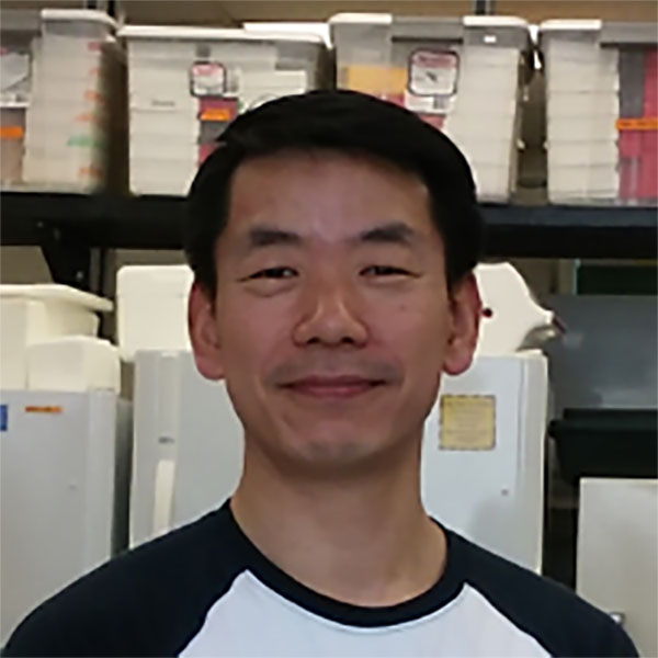 Headshot of Yongsig Kim in a lab with a refrigerator and large storage containers in the background.