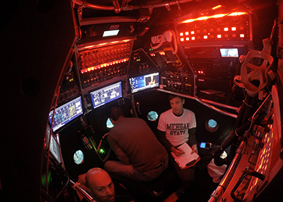 Dalton Hardisty sits with two colleagues in a small, dark chamber — the interior of a submersible vehicle — illuminated by glowing red lights from control panels. 