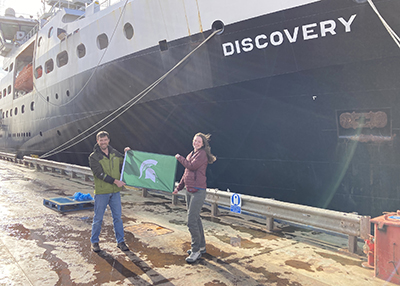 Dalton Hardisty and Kirsten Fentzke hold a green flag with a white Spartan helmet in front of a large research vessel. The ship’s name, “Discovery,” is written in white letters on its dark hull.