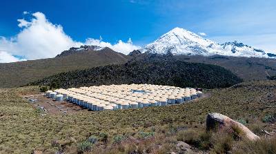 A photo of the High-Altitude Water Cherenkov Observatory in Mexico, showing a grid of 300 water tanks with steel walls and tan caps in the foreground and mountains with blue sky and clouds in the background.