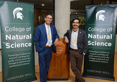 Danny Caballero (right) and Aman Yadav pose together in front of two College of Natural Science banners for a photo following their investiture ceremony. 