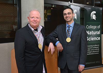Bjoern Hamberger (left) and Ben Orlando pose with their investiture medallions up by the podium with a College of Natural Science banner in the background after their investiture ceremony. Harley J. Seeley