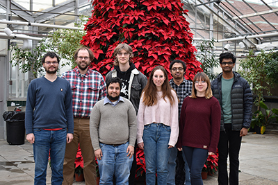 MSU postdoctoral researcher Martin Kulke (far left) and Assistant Professor Josh Vermaas (second from left) standing in a greenhouse in front of a giant poinsettia tree with six other members of the Vermaas lab. 