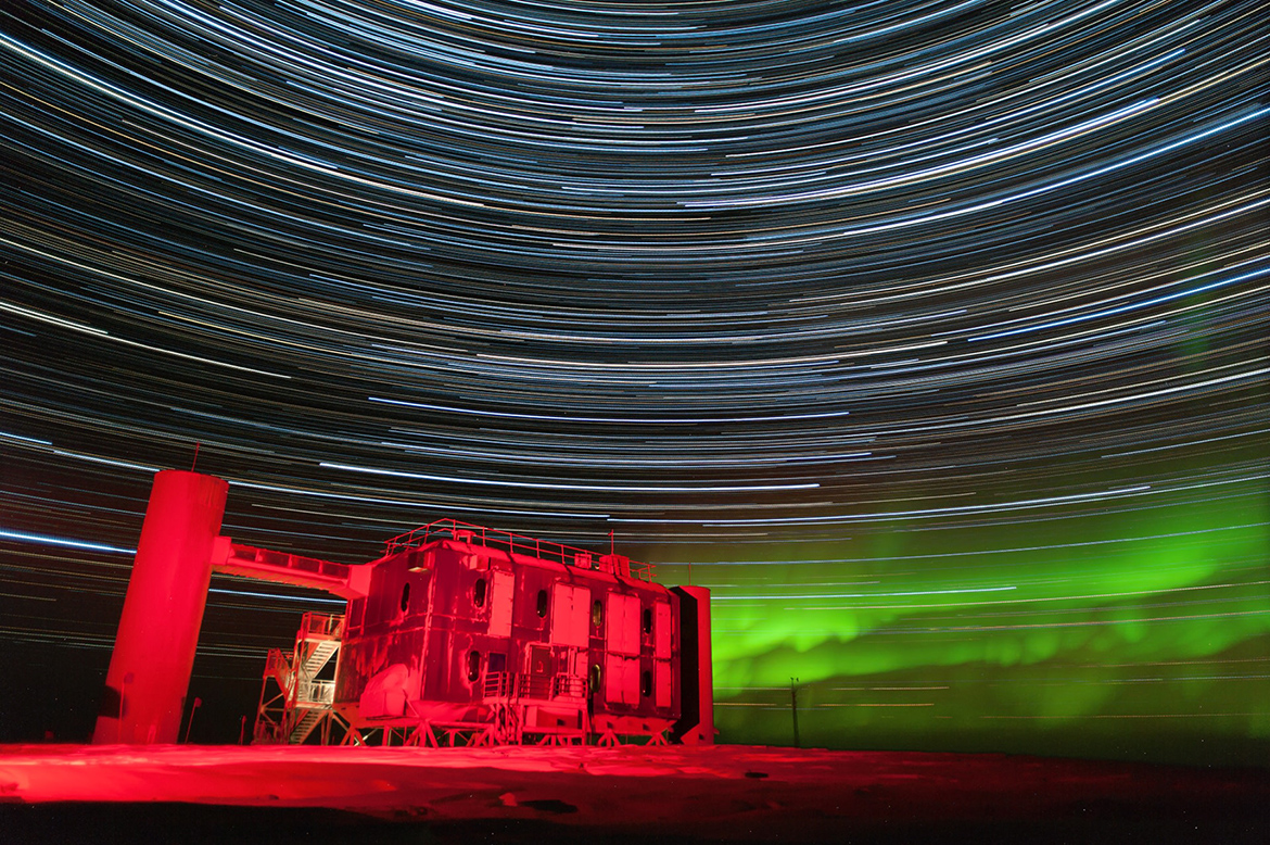The IceCube Lab — a stout, boxy building — stands on Antarctic snow and ice, illuminated by red light against a night sky filled with a green aurora along with blue and white star trails, arching streaks created by starlight and a long exposure setting on the photographer’s camera.