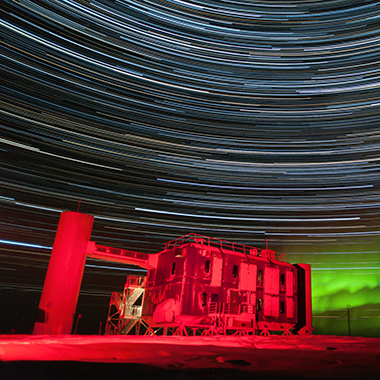 The IceCube Lab — a stout, boxy building — stands on Antarctic snow and ice, illuminated by red light against a night sky filled with a green aurora along with blue and white star trails, arching streaks created by starlight and a long exposure setting on the photographer’s camera.