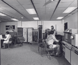 Historic, black and white photo featuring three mass spec users. A handful of machines line the wall of the carpeted space. One user sits at a computer console, one at a mass spec instrument, while another stands. The nearest user operating a mass spec instrument is dressed for the era: boots, wide-bottomed white pants, and a white dress shirt with a decorative pattern on its back.