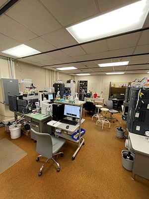 Mass spec instruments, computers, tables, stools, and chairs crowd the original facility space. The room is framed with low paneled ceilings and orange-brown carpet. Wires and containers rest on most surfaces. 