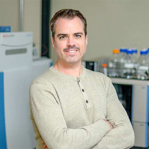 MSU researcher Robert Quinn poses for a waist-up portrait. He wears a tan Henley sweater with arms folded. Behind him is an out-of-focus laboratory space: a large instrument and glass containers. 