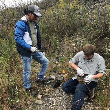 Two men work, both wearing white rubber gloves, on a rocky slope with sparse vegetation. One is standing and holding a trowel full of dirt. The other is sitting and putting dirt into a metal bowl on his lap. A spray bottle lies on the ground nearby. 