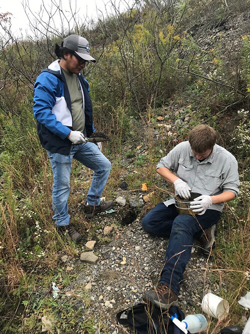 Two men work, both wearing white rubber gloves, on a rocky slope with sparse vegetation. One is standing and holding a trowel full of dirt. The other is sitting and putting dirt into a metal bowl on his lap. A spray bottle lies on the ground nearby. 