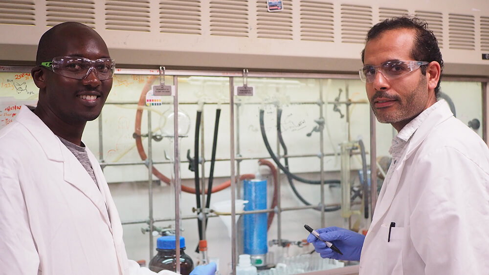 herif Ramadan (right) and Morgan Mayieka (left) stand in front of fume hood with notes and molecular structures sketched on its clear sash.