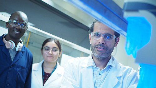 Sherif Ramadan, Ananya Mukherji and Morgan Mayieka (right to left) stand in front of a fume hood in a chemistry lab. Inside the fume hood, clear sample tubes are illuminated with a bright blue light.