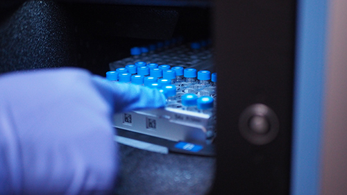 A purple-gloved hand points at a tray filled with small vials with blue caps sits inside a small opening in a mass spectrometer.