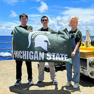 Fan Wang, S. Shawn Wei and Yurong Zhang (left to right) stand on the deck of U.S. Research Vessel Thomas G. Thompson holding a green flag with a white Spartan helmet that says “Michigan State.” White clouds spread across a blue sky and sea in the background