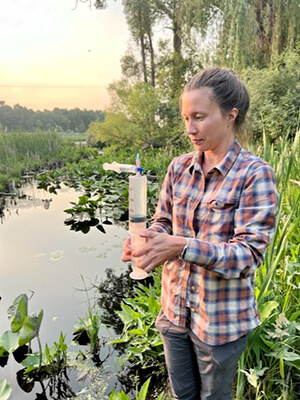 Kelly Aho measures a sample of water in a graduated cylinder in a stream bordered by green trees and grasses 