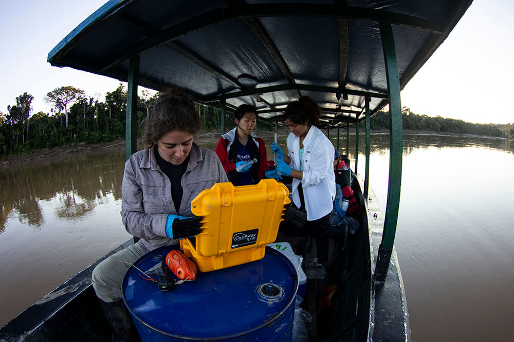Jackie Gerson (front) and two Duke undergraduate students ride on a river boat, looking at a sample in a yellow hardcase resting on a blue barrel..