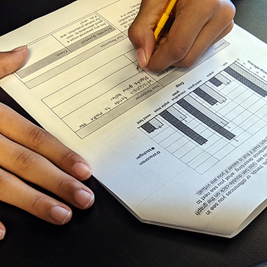 A close-up of a student’s hands filling out a blank table on a worksheet underneath a bar graph comparing Michigan and Wisconsin crop yields.