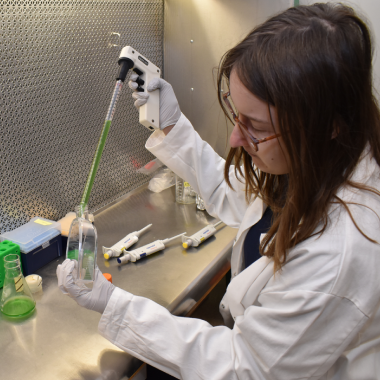 A person in a lab coat uses a pipette to move green liquid to a scientific flask.