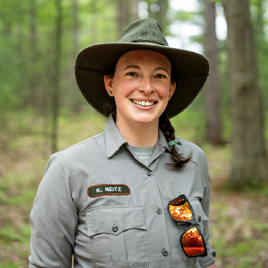 A woman in a park ranger outfit stands in the woods.