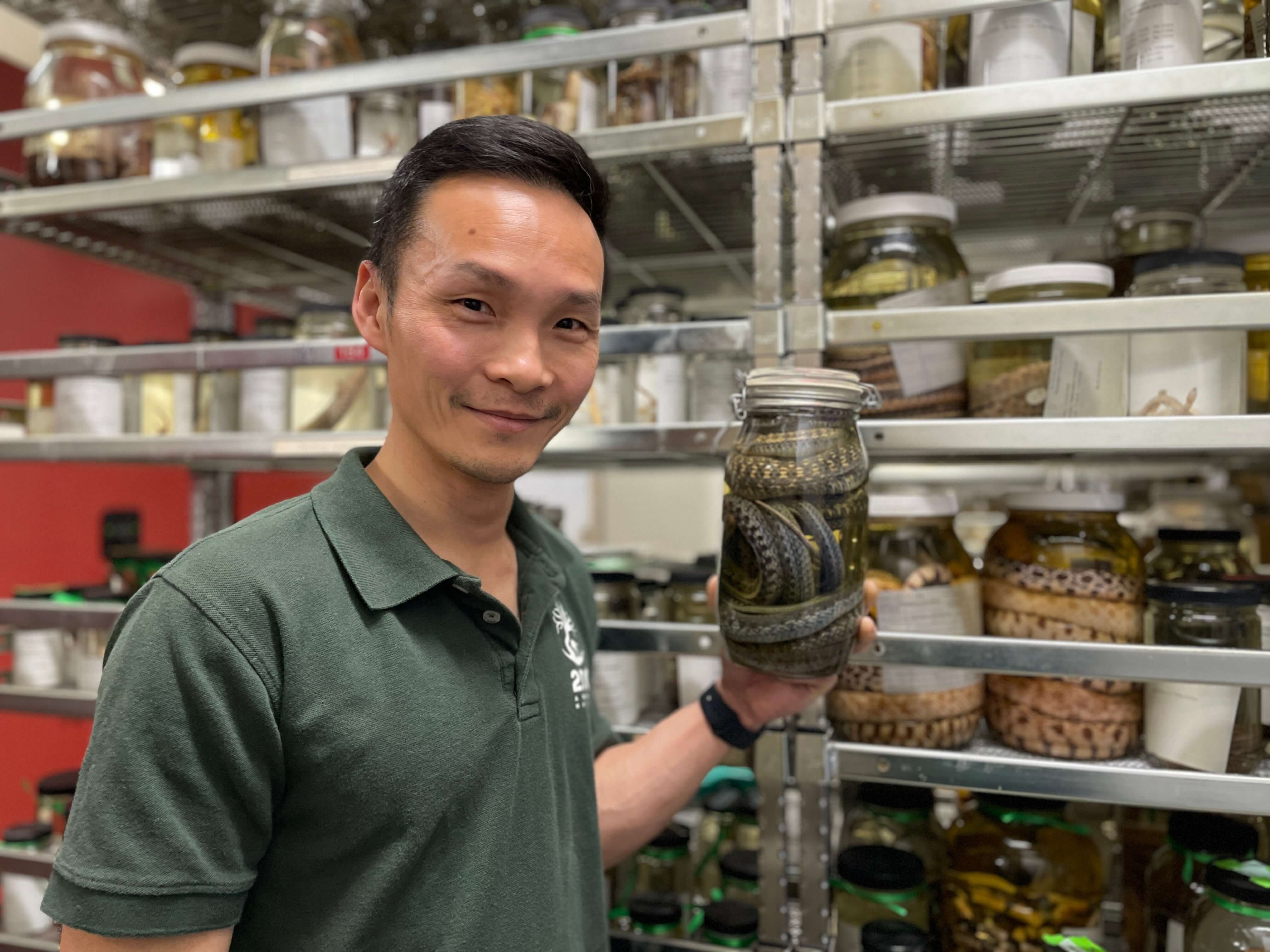A man holding a preserved snake in a jar smiles at the camera. Behind him, multiple other preserved animal specimens can be found.