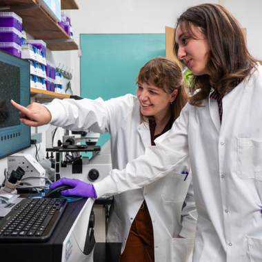 Two researchers in lab coats stand beside a computerized microscope.