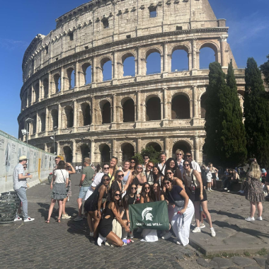 A group of college students with an MSU flag pose in front of the Colosseum.