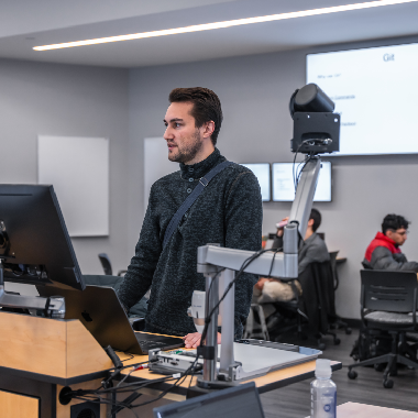 A teacher stands in a classroom by a projector.