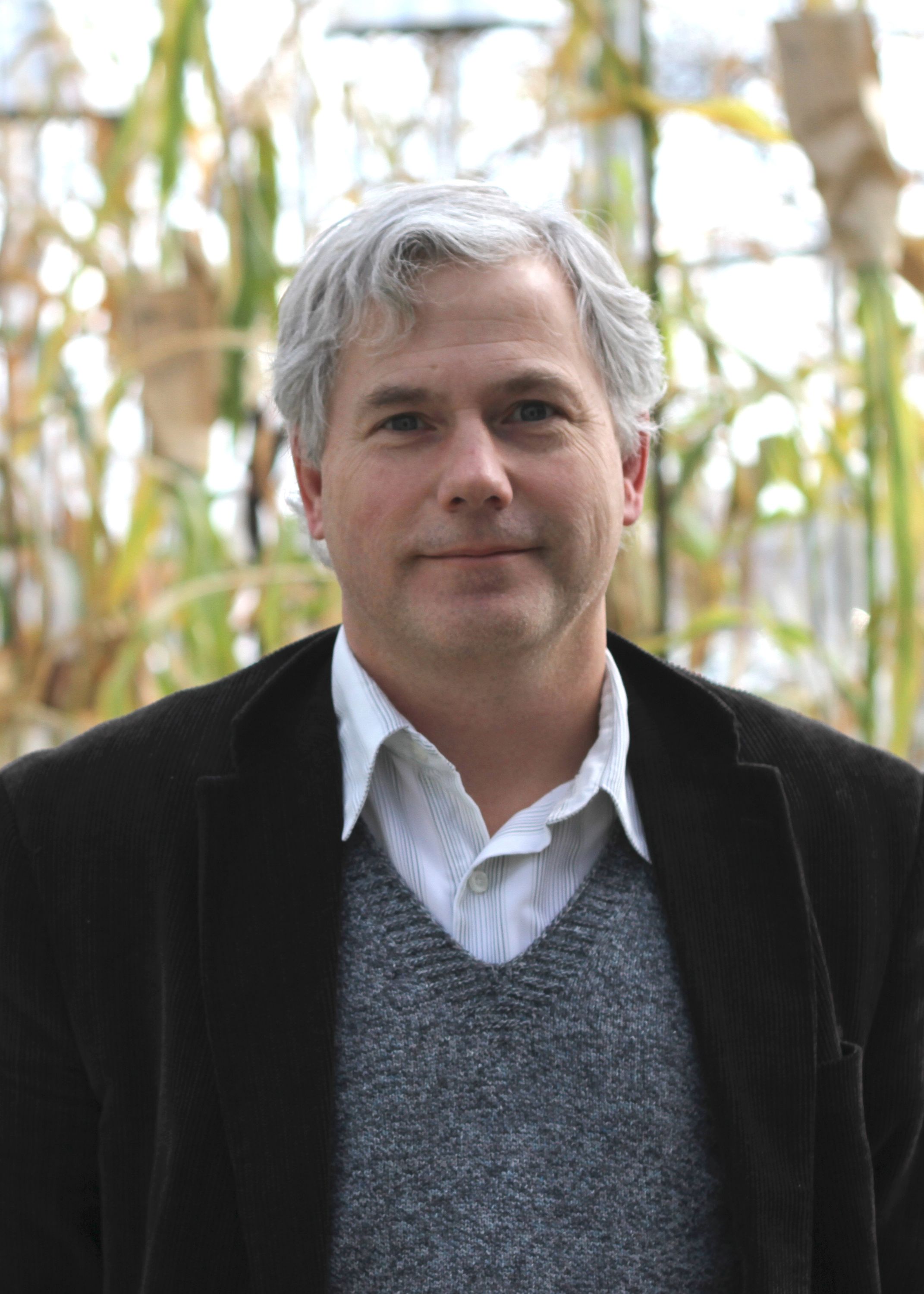 Professor Erich Grotewold in suite coat, sweater, and dress shirt posing in front of plants in greenhouse.