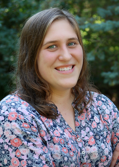 Elizabeth Heath-Heckman smiles at the camera. She is wearing a floral shirt and is standing in front of some trees.