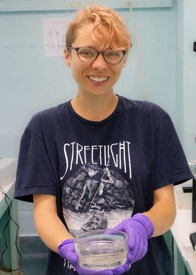Doctoral student Sam Westcott smiles at the camera. She is holding a dish with newly hatched newts in it.