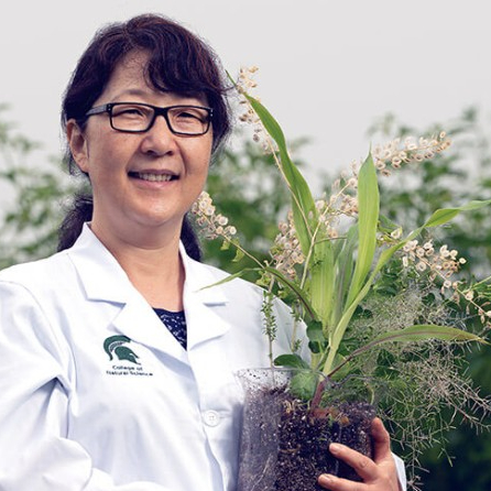 portrait of MSU researcher Sue Rhee wearing a white lab coat and holding a plant that is growing from a large glass beaker