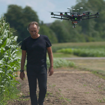 Bruno Basso walking on the edge of a corn field.  A drone is flying over his left shoulder.