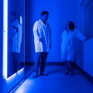 Two figures stand beside a large, blue-lit microscope tower.