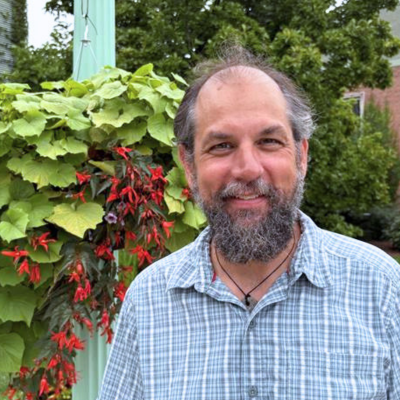 David Tank stands next to a hanging plant with red flowers