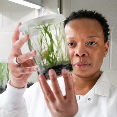 black woman wearing a lab coat holds up a container filled with plants and is inspecting them through the light.