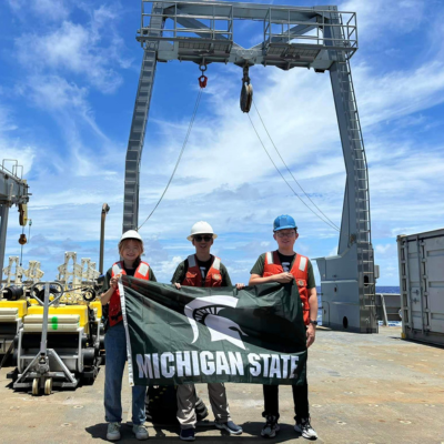 three people standing on a ship holding a Michigan State University flag with a Spartan Helmet