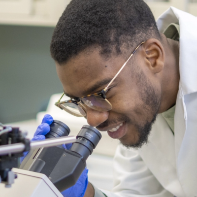 man wearing lab coat and blue gloves looks into a microscope