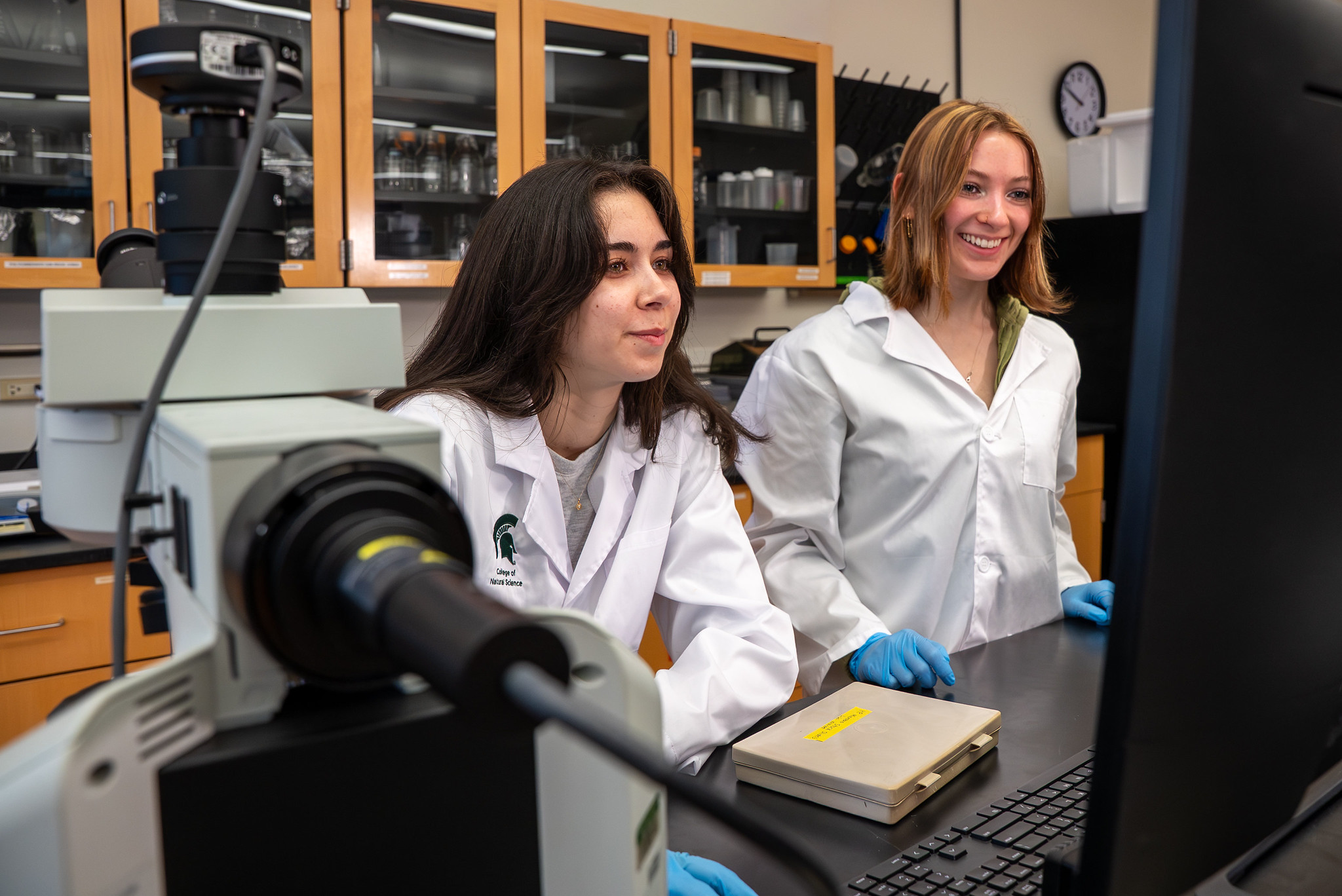 Two students in white labcoats working on a computer