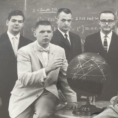 four white male college students are gathered around a globe that has chalk marks. This is a black and white photo from 1960.