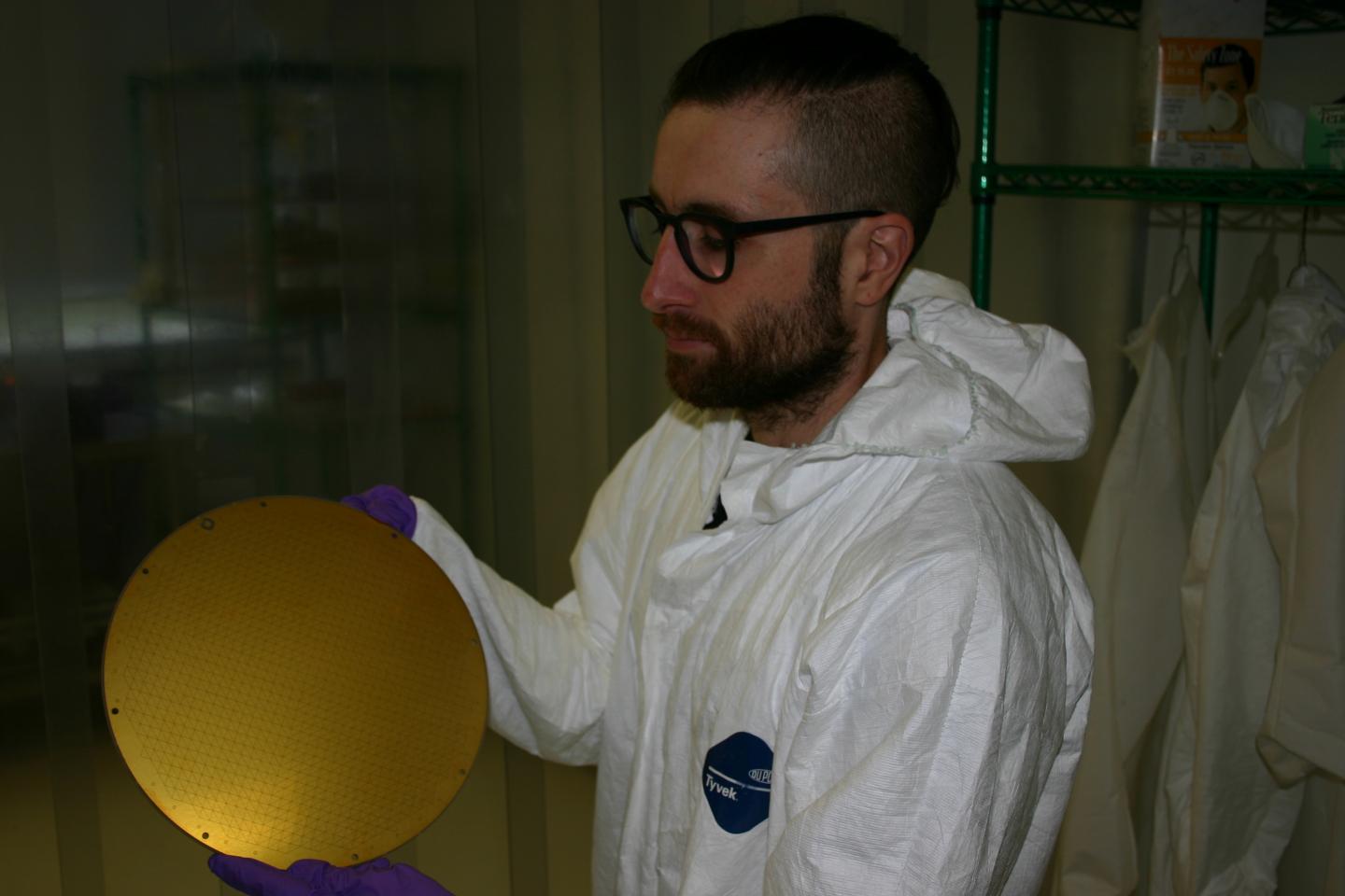 Yassid Ayyad wears a white cleanroom suit and holds a gold-colored disc, about the size of a soccer ball.